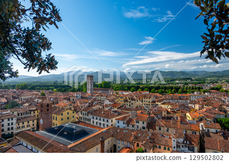 Panoramic view of Lucca, Italy, with red brick rooftops and historic churches seen from above. Tuscan hills and mountains form a stunning summer backdrop under a clear blue sky. Panoramic view of Lucca, Italy, with red brick rooftops and historic churches seen from above. Tuscan hills and mountains form a stunning summer backdrop under a clear blue sky. 129502802