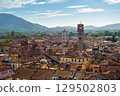 Panoramic view of Lucca, Italy, with red brick rooftops and historic churches seen from above. Tuscan hills and mountains form a stunning summer backdrop under a clear blue sky. 129502803