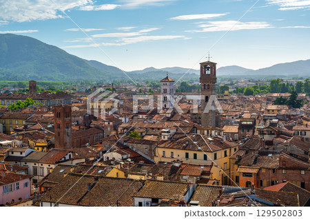Panoramic view of Lucca, Italy, with red brick rooftops and historic churches seen from above. Tuscan hills and mountains form a stunning summer backdrop under a clear blue sky. Panoramic view of Lucca, Italy, with red brick rooftops and historic churches seen from above. Tuscan hills and mountains form a stunning summer backdrop under a clear blue sky. 129502803
