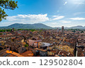 Panoramic view of Lucca, Italy, with red brick rooftops and historic churches seen from above. Tuscan hills and mountains form a stunning summer backdrop under a clear blue sky. 129502804