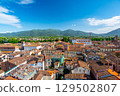 Panoramic view of Lucca, Italy, with red brick rooftops and historic churches seen from above. Tuscan hills and mountains form a stunning summer backdrop under a clear blue sky. 129502807