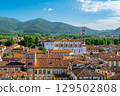 Panoramic view of Lucca, Italy, with red brick rooftops and historic churches seen from above. Tuscan hills and mountains form a stunning summer backdrop under a clear blue sky. 129502808