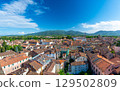 Panoramic view of Lucca, Italy, with red brick rooftops and historic churches seen from above. Tuscan hills and mountains form a stunning summer backdrop under a clear blue sky. 129502809