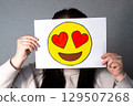 Woman holding heart eyed emoji mask in front of her face. Social media trends, Gen Z marketing, World emoji day 17 July. 129507268