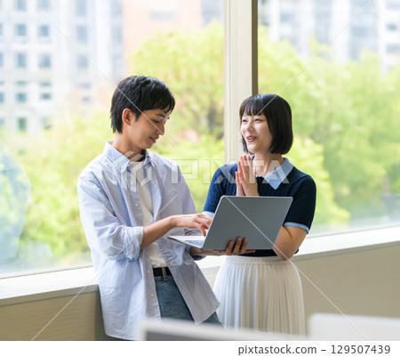 Male and female university students having a conversation by the window with a laptop Male and female university students having a conversation by the window with a laptop 129507439