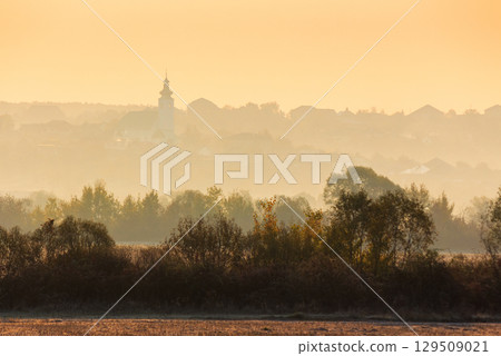 rural landscape on an autumn morning. row of trees along the empty fields. village with church on the hill behind the forest in the hazy distance 129509021
