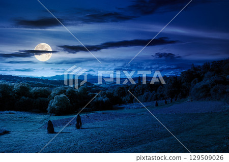 countryside landscape on september night. village in the valley and rural fields on the forested hills in full moon light. mountain ridge with high peak in the distance countryside landscape on september night. village in the valley and rural fields on the forested hills in full moon light. mountain ridge with high peak in the distance 129509026