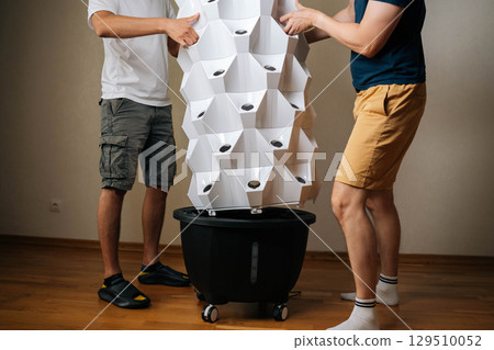 Cropped shot of two urban farmers placing main body of vertical hydroponic tower garden onto its base, working together to set up indoor space for cultivating fresh plants. Concept of home farming. Cropped shot of two urban farmers placing main body of vertical hydroponic tower garden onto its base, working together to set up indoor space for cultivating fresh plants. Concept of home farming. 129510052