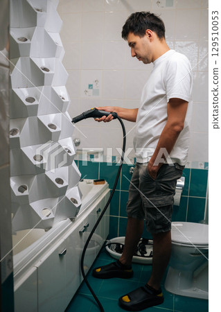 Vertical side view of man using steam cleaner to sanitize vertical hydroponic tower garden in bathroom, demonstrating modern and sustainable approach to indoor gardening. Concept of home gardening. Vertical side view of man using steam cleaner to sanitize vertical hydroponic tower garden in bathroom, demonstrating modern and sustainable approach to indoor gardening. Concept of home gardening. 129510053