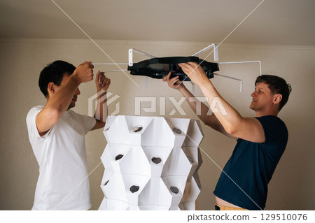 Two agricultural technician assembling indoor vertical hydroponic tower garden, placing top part with grow light onto structure, ready to start growing fresh produce at home. Concept of indoor farming Two agricultural technician assembling indoor vertical hydroponic tower garden, placing top part with grow light onto structure, ready to start growing fresh produce at home. Concept of indoor farming 129510076