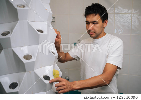 Gardener male wiping vertical hydroponic tower garden with yellow sponge, maintaining clean growing environment in contemporary bathroom space. Concept of healthy and sustainable cultivation. Gardener male wiping vertical hydroponic tower garden with yellow sponge, maintaining clean growing environment in contemporary bathroom space. Concept of healthy and sustainable cultivation. 129510096