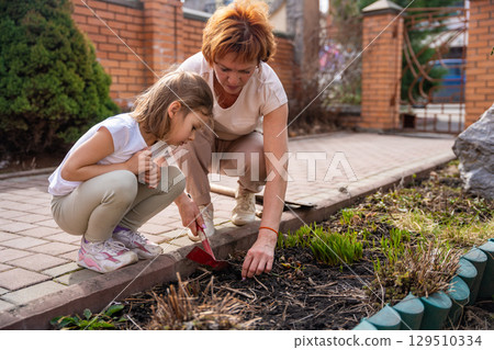 Woman gardening with a young girl in the backyard, teaching her how to care for flowers. Family bonding, outdoor learning, and eco-friendly lifestyle. 129510334