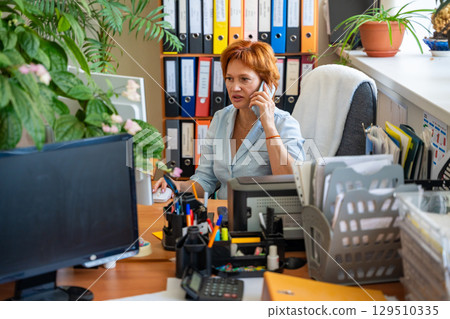 Woman sitting at her desk in the office, talking on the phone while working on the computer. Business communication, multitasking, and everyday coordination at the workplace. 129510335