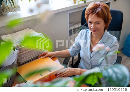 Woman working with folders and documents at her desk in an office setting. Paperwork routine, archive management, and administrative desk tasks. 129510336