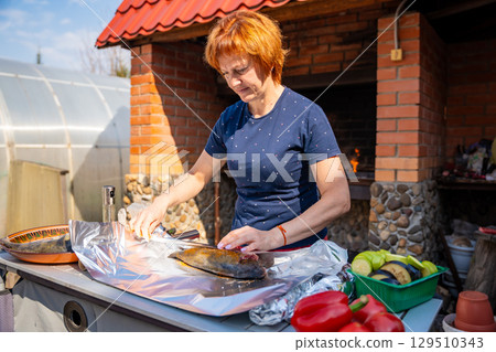 Woman seasoning fish and wrapping it in foil for outdoor grilling in her backyard. Homemade cooking, natural ingredients, and traditional barbecue preparation. 129510343