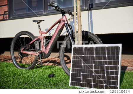 Pink electric mountain bike parked on grassy lawn, connected to solar panel for charging. Bike features rugged tires and positioned beside building, with its shadow cast on wall. Pink electric mountain bike parked on grassy lawn, connected to solar panel for charging. Bike features rugged tires and positioned beside building, with its shadow cast on wall. 129510424