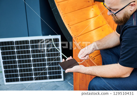 Cropped view of man connecting smartphone to photovoltaic solar panel. Integration of sustainable renewable energy into everyday life, demonstrating practical use of solar power for charging devices. 129510431