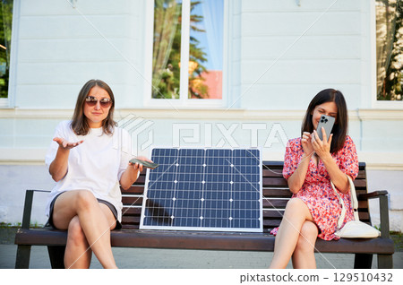 Young girl can't charge her phone because her friend used all energy. Confused girl doesn't know how to charge phone from solar battery. Two female sitting on bench with solar panel between them. Young girl can't charge her phone because her friend used all energy. Confused girl doesn't know how to charge phone from solar battery. Two female sitting on bench with solar panel between them. 129510432