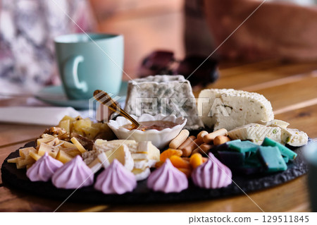 Cheese platter with various cheeses, sweets, and a cup of tea on a wooden table in a cozy setting 129511845