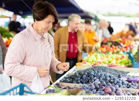 Positive mature woman in casual clothes picking fresh plums during shopping at grocery market 129512220