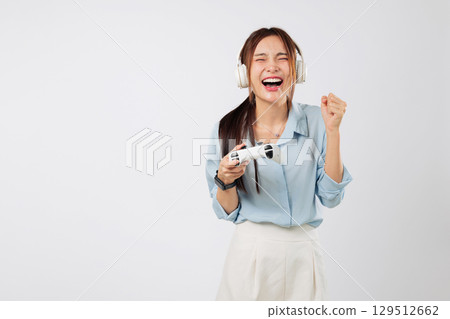Game action captured as teen woman raises fist in victory, headphones on, eyes on screen, gripping game controller while smiling proudly. isolated studio white background Game action captured as teen woman raises fist in victory, headphones on, eyes on screen, gripping game controller while smiling proudly. isolated studio white background 129512662