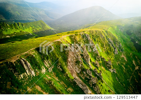 Breathtaking aerial view of lush, green mountain ridge with rugged rocky cliffs. Landscape extends into misty valleys and distant mountains, bathed in soft, golden sunlight. Carpathians, Chornogora. Breathtaking aerial view of lush, green mountain ridge with rugged rocky cliffs. Landscape extends into misty valleys and distant mountains, bathed in soft, golden sunlight. Carpathians, Chornogora. 129513447