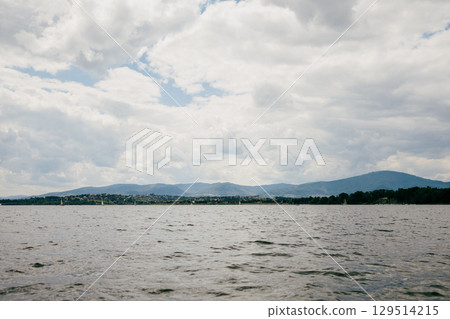 Lake reflecting cloudy sky with distant mountains and sailboats, offers serene view perfect for enjoying natura tranquility. Cloudy sky reflecting on lake, revealing distant mountains and sailboats Lake reflecting cloudy sky with distant mountains and sailboats, offers serene view perfect for enjoying natura tranquility. Cloudy sky reflecting on lake, revealing distant mountains and sailboats 129514215