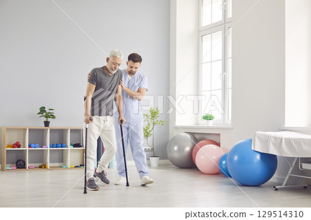 Portrait of young nurse helping a senior patient to walk with his crutches in rehabilitation clinic Portrait of young nurse helping a senior patient to walk with his crutches in rehabilitation clinic 129514310