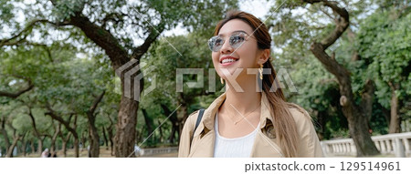 Young woman smiling while walking through a park on a sunny autumn day, showcasing a stylish trench coat and sunglasses 129514961