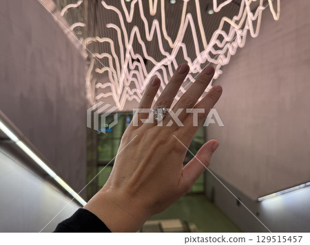 Landscape close up photo of a woman's hand wearing a large oval cut diamond engagement ring, held up against a modern escalator with artistic neon lights in the background. Landscape close up photo of a woman's hand wearing a large oval cut diamond engagement ring, held up against a modern escalator with artistic neon lights in the background. 129515457