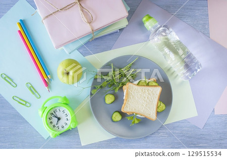 Back to school concept. On the table is a school lunch, an alarm clock and stationery. A sandwich, an apple, a bottle of water and textbooks. Back to school concept. On the table is a school lunch, an alarm clock and stationery. A sandwich, an apple, a bottle of water and textbooks. 129515534