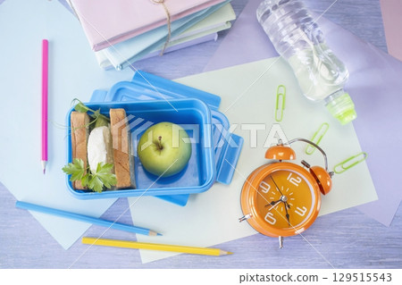 Back to school concept, school food. Lunch box with healthy sandwich, alarm clock, stack of books, apple and bottle of water on chalkboard background. Back to school concept, school food. Lunch box with healthy sandwich, alarm clock, stack of books, apple and bottle of water on chalkboard background. 129515543