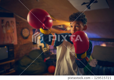 Teen boy wearing red boxing gloves practices punches in cozy attic with retro posters. 129515721