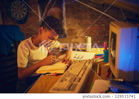 Focused boy writing in notebook at desk with vintage computer 129515741