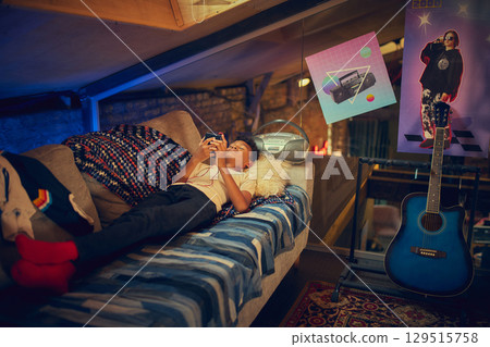Boy lying on couch in retro-decorated attic playing handheld console 129515758
