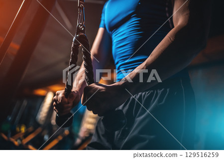 Closeup male bodybuilder performing triceps pushdown with rope in gym, Cropped shot man training arm muscles on cable machine during workout 129516259