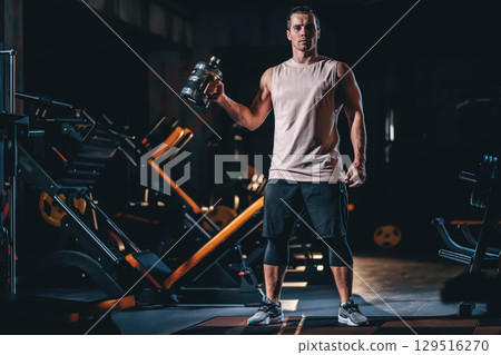 Male bodybuilder holding sports drink with BCAA for recovery in gym, Man standing in sport gym with creatine and protein drink after workout Male bodybuilder holding sports drink with BCAA for recovery in gym, Man standing in sport gym with creatine and protein drink after workout 129516270