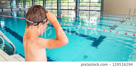 Smiling boy in swimming goggles sitting by indoor pool ready to have fun in water. Concept of kids sport, habits 129516294