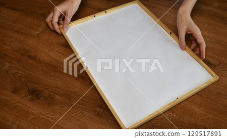 Female hands hold sheet of white paper to put it into minimal wooden frame 129517891