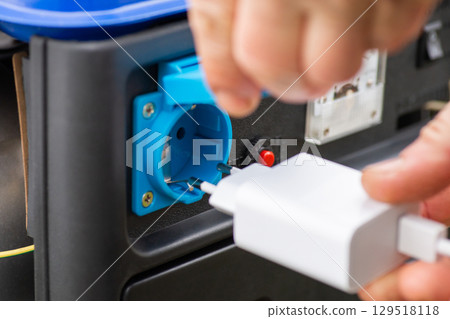 Put on phone charger into the outlet of generator, which produces electricity. Close up of male hand with white plug, concept of charging devices 129518118