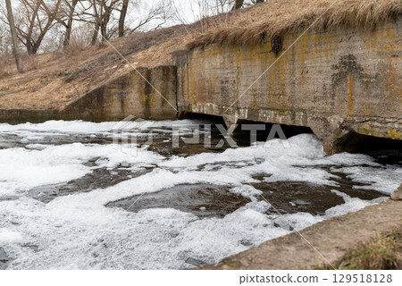 Dam on a small river, a lot of white foam on the water surface. Concept of dirty infected water in the stream, pollution of environment, danger for nature and people 129518128