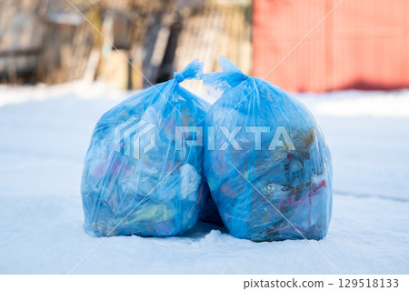 Two blue plastic bags full of garbage on snow, close up. Bags with rubbish on the roadside in winter, concept of recycling and public utilities 129518133