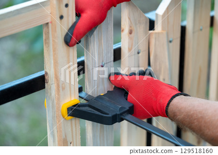 Woodworking sash clamp used to hold planks in place, making new fence. Close up view of man`s hands holding woodworking sash while constructing fence, handiwork concept 129518160