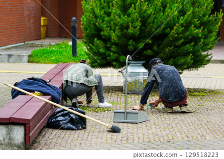 Two people cleaning weeds from pavement in public area with broom, dustpan, and trash bag Two people cleaning weeds from pavement in public area with broom, dustpan, and trash bag 129518223