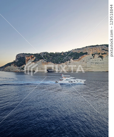 Vibrant evening seascape with a boat cruising away on the blue sea, near the Pirate Cave in Kemer at sunset. Beautiful Turkish mediterranean coastline lined with tree covered rocky cliffs at dusk 129518844