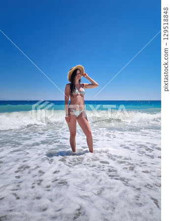 Beautiful woman posing in bikini and straw sun hat, stands in the foamy waves of the clear blue sea, under a cloudless summer sky. Summertime vacation on a tropical beach, relaxation vibes 129518848