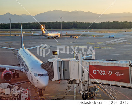 ANTALYA, TURKEY - JULY 5, 2025 Antalya airport scene at sunset and a plane docked at the jet bridge with other aircrafts visible on the tarmac. Distant mountains range silhouette in the background ANTALYA, TURKEY - JULY 5, 2025 Antalya airport scene at sunset and a plane docked at the jet bridge with other aircrafts visible on the tarmac. Distant mountains range silhouette in the background 129518862