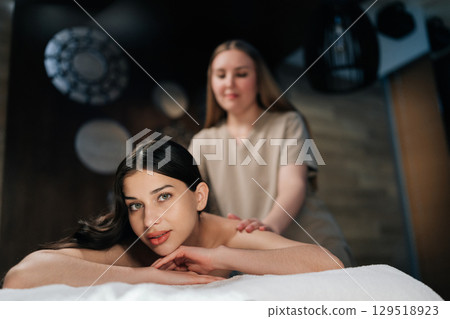 Portrait of pretty brunette woman lying on massage table during professional relaxing massage at spa salon, serene looking at camera. Professional massage therapist performing back treatment. Portrait of pretty brunette woman lying on massage table during professional relaxing massage at spa salon, serene looking at camera. Professional massage therapist performing back treatment. 129518923