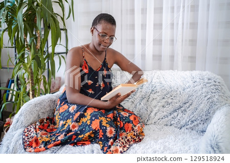 Smiling Black Woman Reading a Book on a Cozy Sofa at Home Smiling Black Woman Reading a Book on a Cozy Sofa at Home 129518924