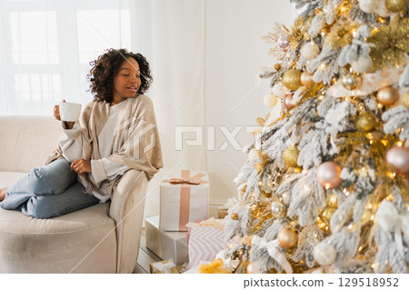 Merry Christmas. African American woman with cup of hot drink coffee tea sitting near Christmas tree at home. Girl in living room with Christmas tree. Christmas hygge holidays at home 129518952